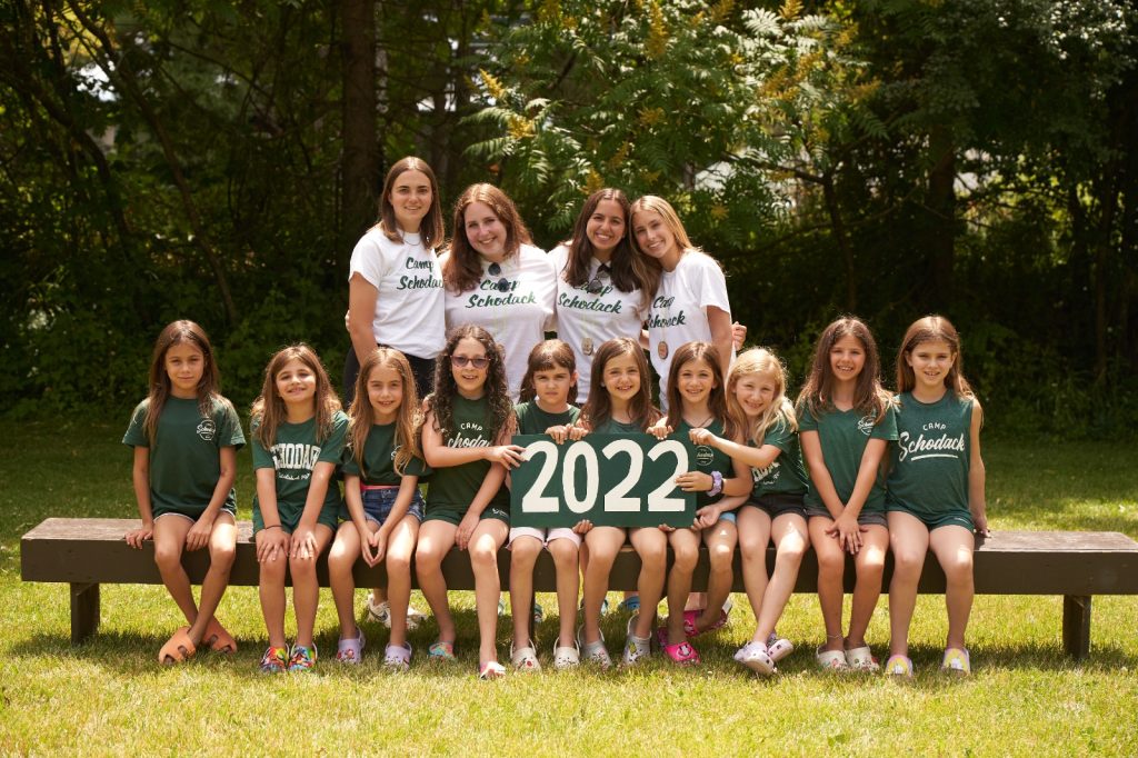 Group of campers and their counselors gathered for a photo. They are seated on a bench and a forest is in the background.