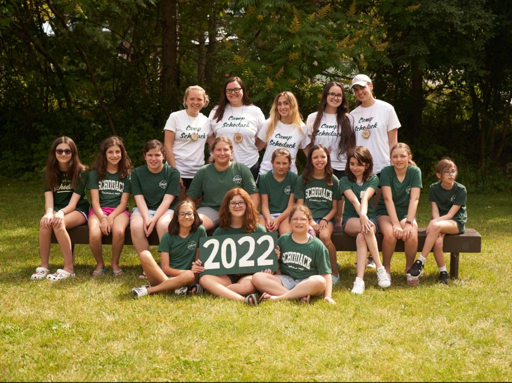 Group of campers and their counselors gathered for a photo. They are seated on a bench and a forest is in the background.