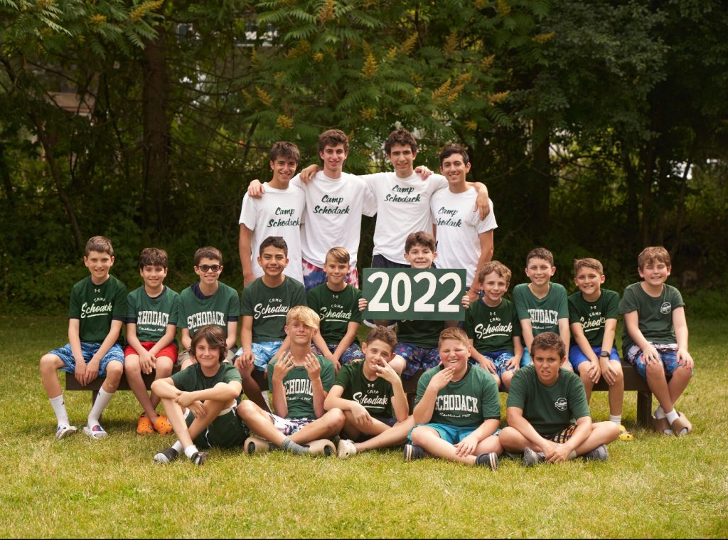 Group of campers and their counselors gathered for a photo. They are seated on a bench and a forest is in the background.