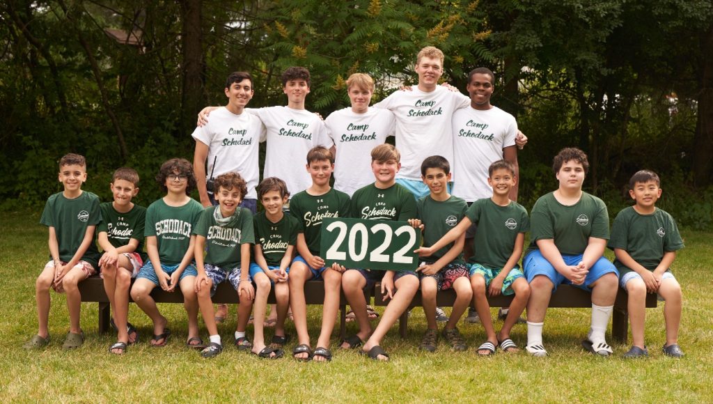 Group of campers and their counselors gathered for a photo. They are seated on a bench and a forest is in the background.