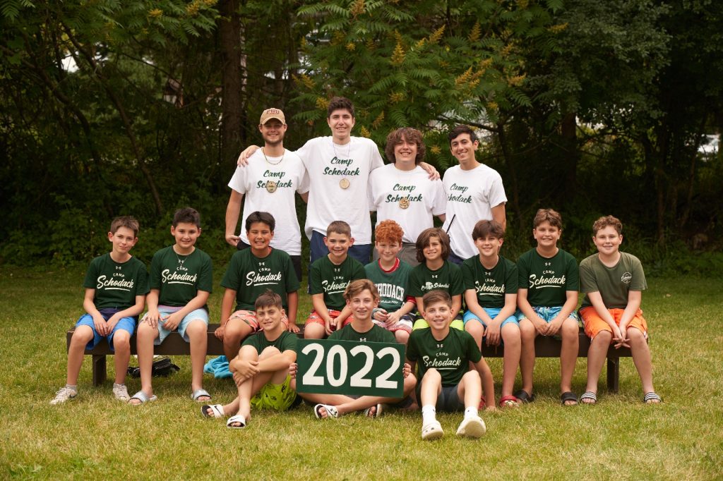 Group of campers and their counselors gathered for a photo. They are seated on a bench and a forest is in the background.