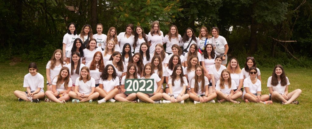 Group of campers and their counselors gathered for a photo. They are seated on a bench and a forest is in the background.