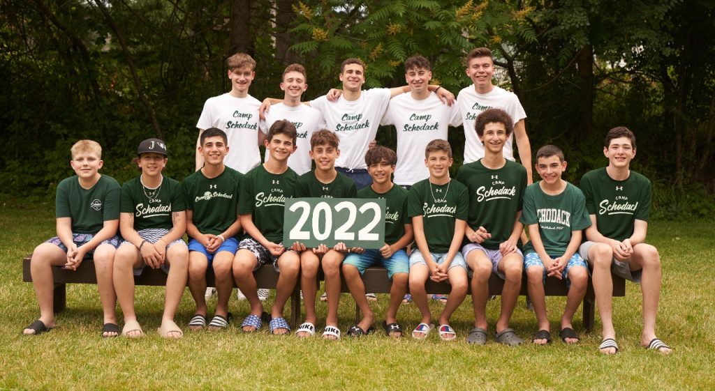 Group of campers and their counselors gathered for a photo. They are seated on a bench and a forest is in the background.