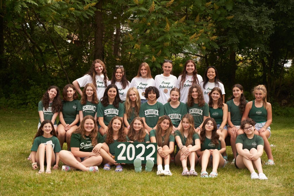 Group of campers and their counselors gathered for a photo. They are seated on a bench and a forest is in the background.