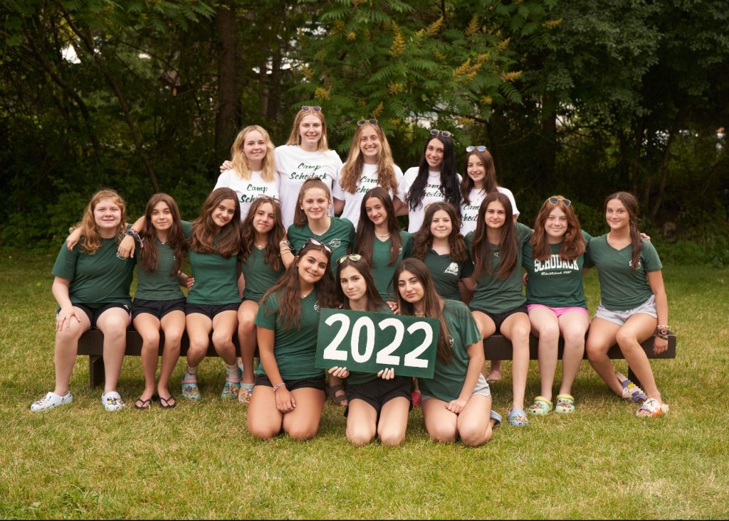 Group of campers and their counselors gathered for a photo. They are seated on a bench and a forest is in the background.