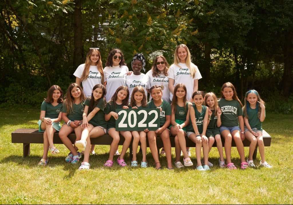 Group of campers and their counselors gathered for a photo. They are seated on a bench and a forest is in the background.