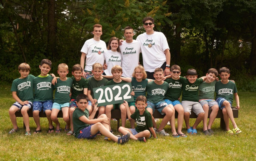 Group of campers and their counselors gathered for a photo. They are seated on a bench and a forest is in the background.