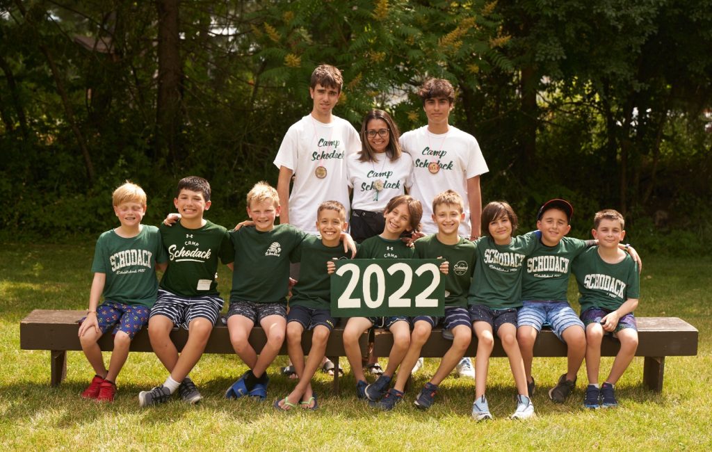 Group of campers and their counselors gathered for a photo. They are seated on a bench and a forest is in the background.