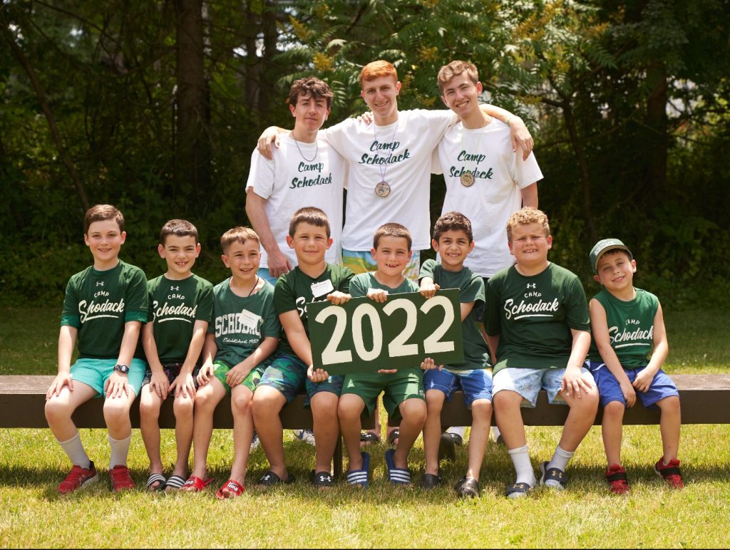 Group of campers and their counselors gathered for a photo. They are seated on a bench and a forest is in the background.