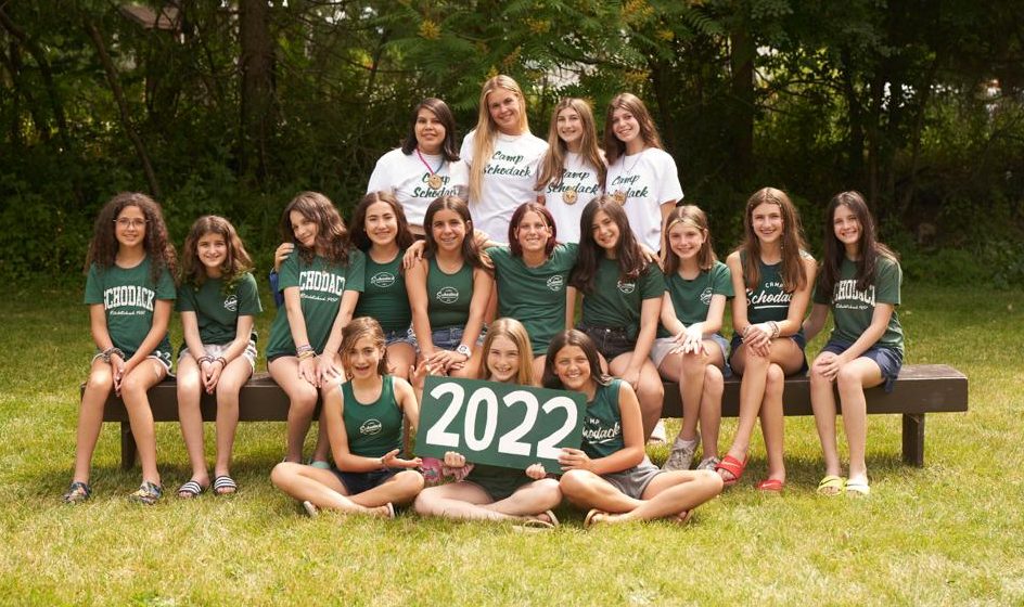 Group of campers and their counselors gathered for a photo. They are seated on a bench and a forest is in the background.