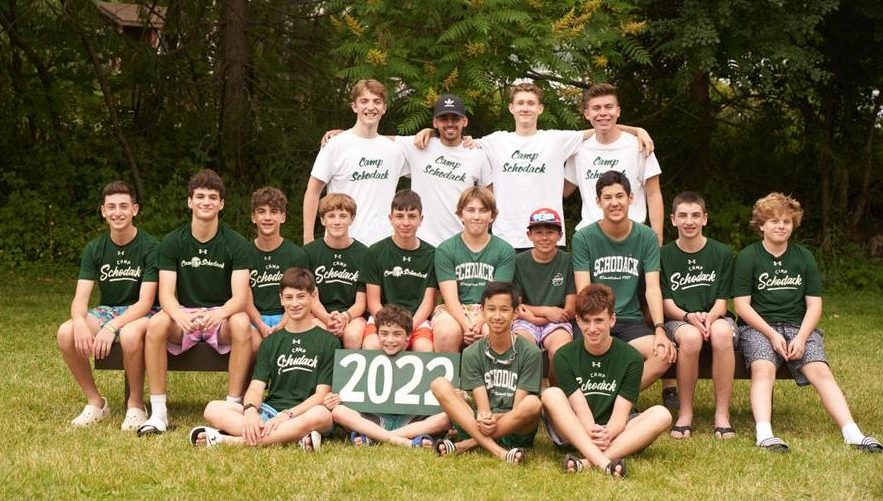 Group of campers and their counselors gathered for a photo. They are seated on a bench and a forest is in the background.