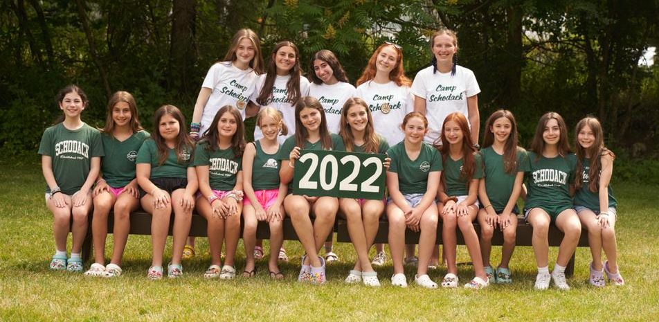 Group of campers and their counselors gathered for a photo. They are seated on a bench and a forest is in the background.